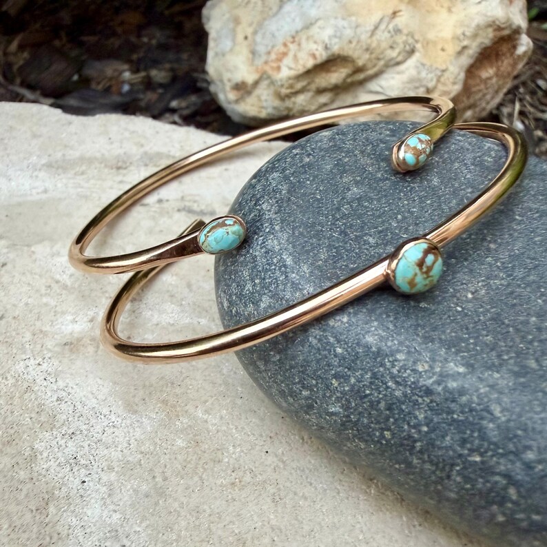 May include: Two gold-colored metal bracelets with turquoise-colored stones. The bracelets are resting on a gray stone. The turquoise stones have brown marbling. The bracelets have an open design.