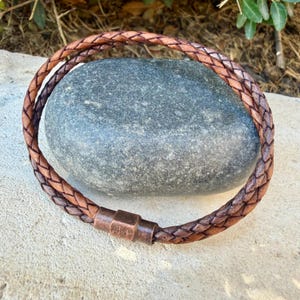 Puede incluir: Una pulsera de cuero trenzado marr&oacute;n con un cierre met&aacute;lico cil&iacute;ndrico de tono cobre. La pulsera est&aacute; formada por m&uacute;ltiples tiras de cuero entrelazadas en un patr&oacute;n circular, creando una apariencia texturizada con variados tonos marrones.