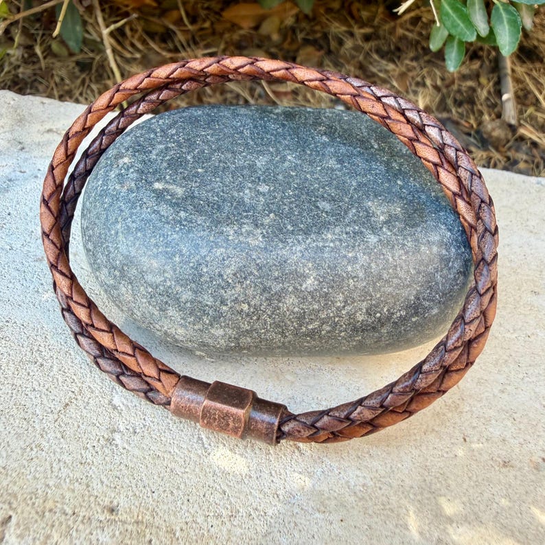 Puede incluir: Una pulsera de cuero trenzado marr&oacute;n con un cierre met&aacute;lico cil&iacute;ndrico de tono cobre. La pulsera est&aacute; formada por m&uacute;ltiples tiras de cuero entrelazadas en un patr&oacute;n circular, creando una apariencia texturizada con variados tonos marrones.