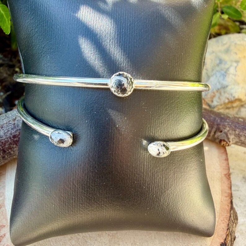 May include: A silver-toned cuff bracelet with a hammered texture and two black stone accents. The open design features a central stone and two end stones. The bracelet is displayed on a black surface.