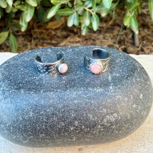 May include: Two open-band rings made of silver-toned metal. The left ring features an etched pattern and a small, round, light pink stone. The right ring has a smooth surface and a larger, round, light pink stone set in a scalloped bezel.
