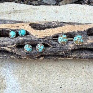 May include: Six pairs of turquoise and brown stud earrings with silver-colored settings are displayed on a piece of driftwood. The earrings have a round shape and a textured appearance, with a mix of blue and brown colors.