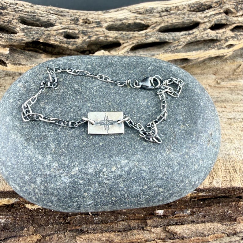 K&ouml;nnte beinhalten: Ein silbernes Kettenarmband mit einem rechteckigen Anh&auml;nger mit einem Kreuzdesign. Das Armband wird auf einem grauen Stein vor einem Hintergrund aus Holz pr&auml;sentiert.