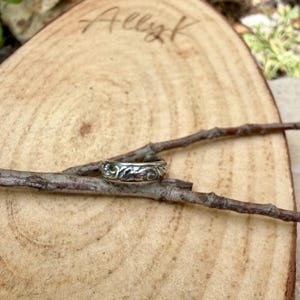 May include: A silver ring with an intricate floral design rests on a small twig. The ring is set against a natural wood background with the handwritten name "Allyk" visible. The ring's detailed pattern is highlighted by the natural lighting.