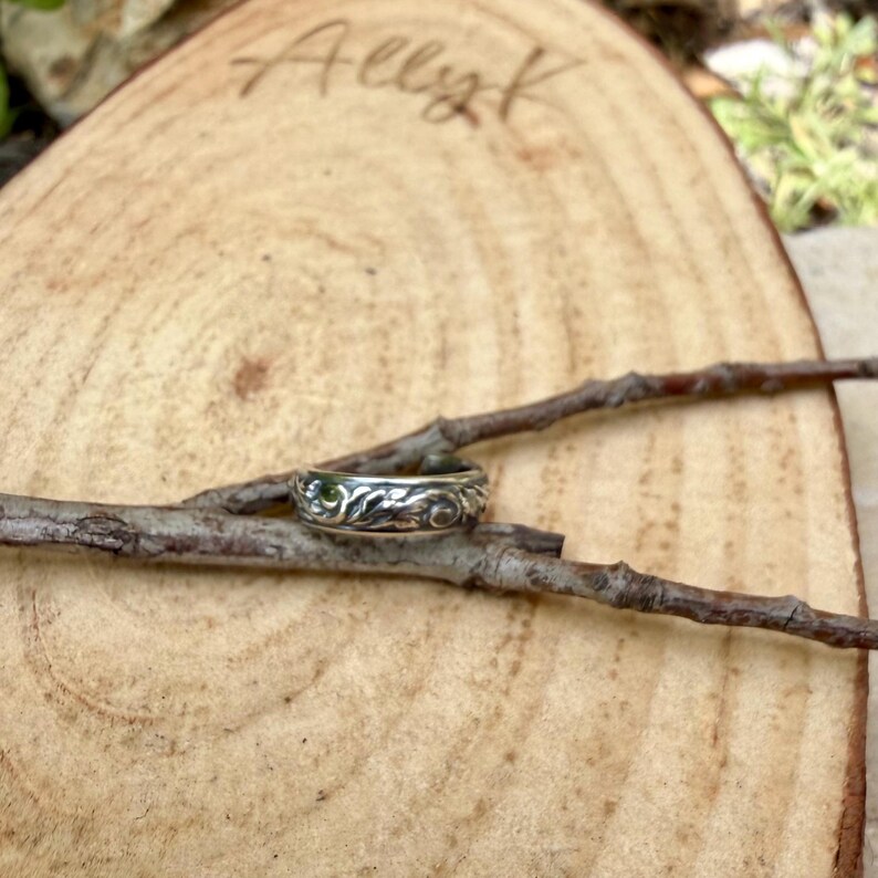 May include: A silver ring with an intricate floral design rests on a small twig. The ring is set against a natural wood background with the handwritten name "Allyk" visible. The ring's detailed pattern is highlighted by the natural lighting.