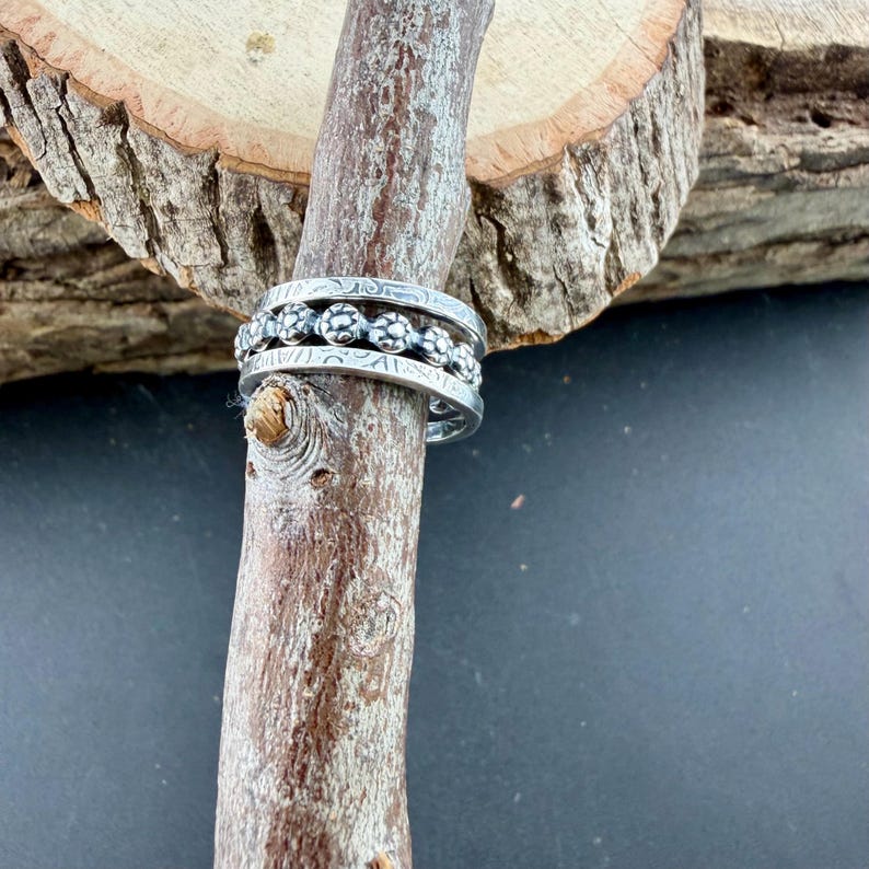 May include: Three silver rings with intricate floral and patterned designs are stacked on a wooden stick. The rings have a vintage or handcrafted appearance, with detailed engravings. The rings are displayed against a natural wood background.