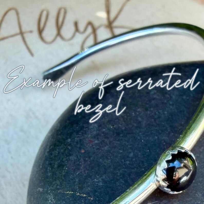 May include: A close-up of a silver-colored bracelet with a black, round stone set in a serrated bezel. The bracelet is resting on a dark, textured surface. Text in the image reads "Example of serrated bezel."