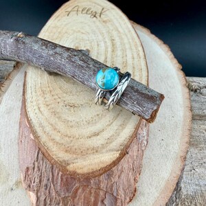 May include: Two silver rings with a turquoise stone and a leaf design, displayed on a wooden branch. The rings are set against a wooden background with the handwritten text "Ally K".