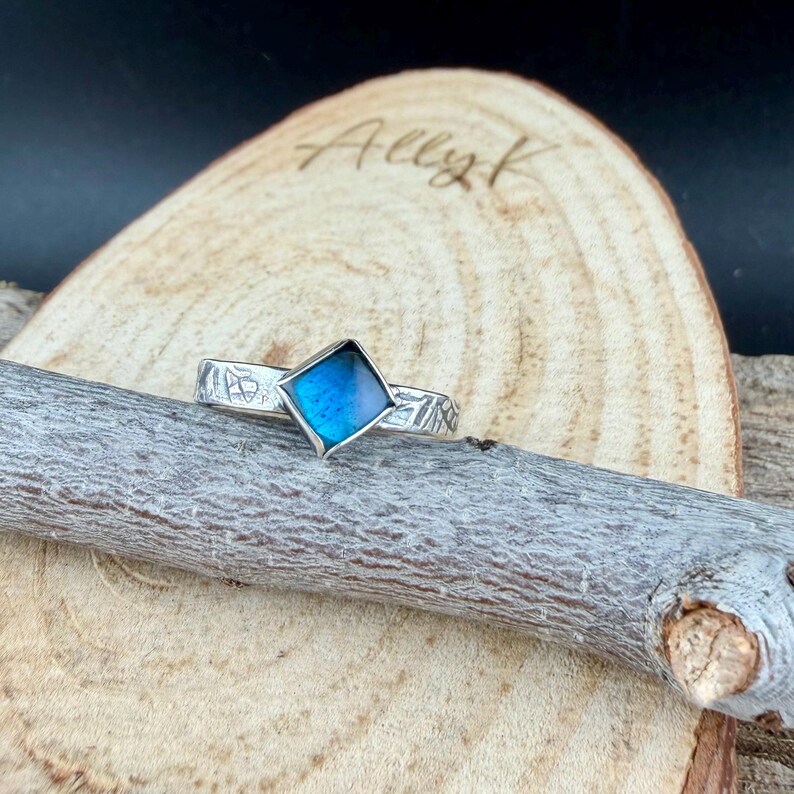 May include: A silver ring with a square, iridescent blue center stone. The band features etched designs. The ring is displayed on a wooden surface with the name "Ally K" written on it.