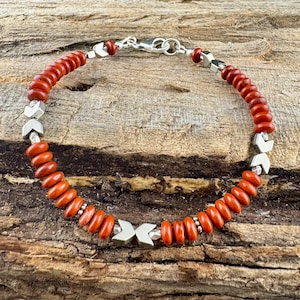 May include: A beaded bracelet featuring reddish-orange beads and silver-colored arrow-shaped accents. The bracelet is strung on a silver chain and has a silver clasp. The bracelet is displayed on a wooden surface.