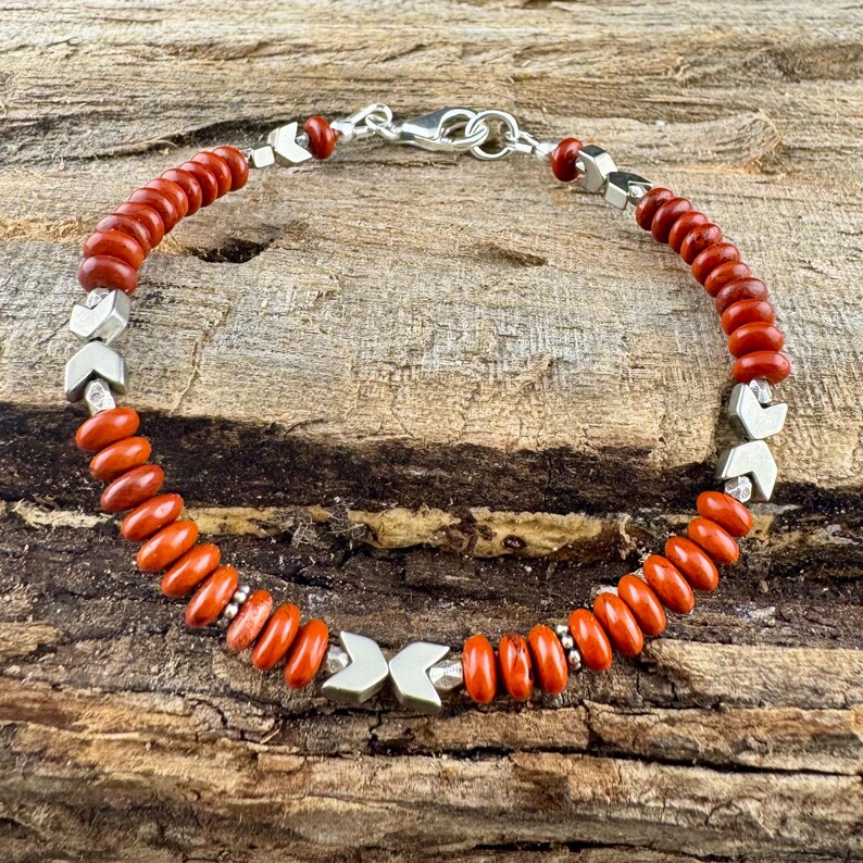 May include: A beaded bracelet featuring reddish-orange beads and silver-colored arrow-shaped accents. The bracelet is strung on a silver chain and has a silver clasp. The bracelet is displayed on a wooden surface.