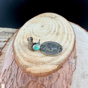 May include: A silver-toned ring with a turquoise stone and a hammered texture, next to a silver coin with a buffalo design. The ring has an open design. The coin reads "FIVE CENTS".