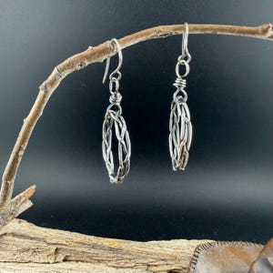 Op de afbeelding: Een paar zilverkleurige draadorbellen met een ovaal kooiontwerp gemaakt van in elkaar gevlochten metalen draden. Elke oorbel hangt aan een eenvoudige haak en heeft een getextureerd, geweven uiterlijk met een gepolijste metalen afwerking.