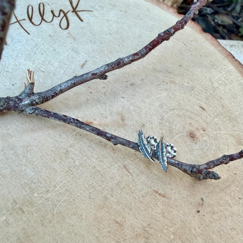 May include: A pair of silver feather earrings displayed on a small tree branch. The earrings have detailed feather designs and are set against a light brown wooden background. The word "allyk" is written in cursive in the upper left corner.