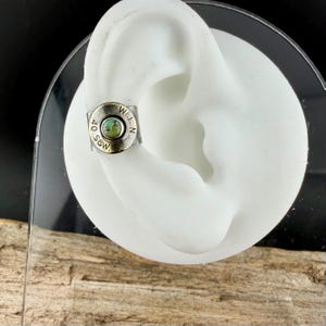 May include: A silver ear cuff featuring a turquoise stone. The cuff is circular with the text "40 S&W" and "W.L.N." engraved around the edge. The cuff is displayed on a white ear model.