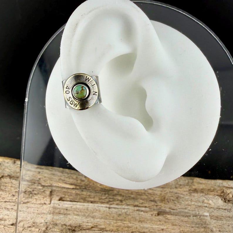 May include: A silver ear cuff featuring a turquoise stone. The cuff is circular with the text "40 S&W" and "W.L.N." engraved around the edge. The cuff is displayed on a white ear model.