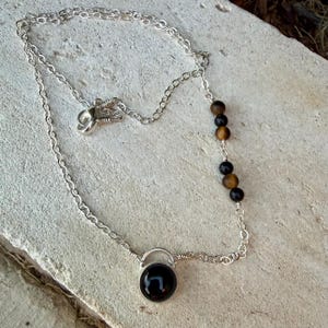 May include: A silver chain necklace featuring a single round black stone pendant at the center and a series of five alternating black and brown spherical beads integrated into one side of the chain, secured with a silver lobster claw clasp.