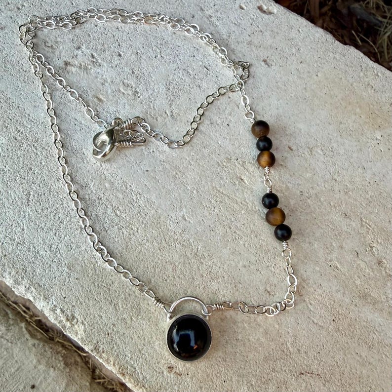 May include: A silver chain necklace featuring a single round black stone pendant at the center and a series of five alternating black and brown spherical beads integrated into one side of the chain, secured with a silver lobster claw clasp.