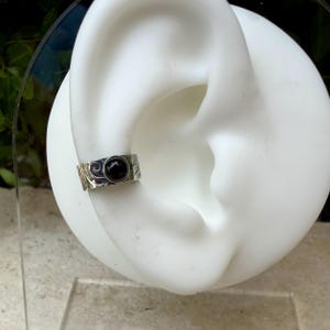 May include: A silver ear cuff with a black stone set in the center. The cuff has a textured surface with a swirling design. The jewelry is displayed on a white ear model.