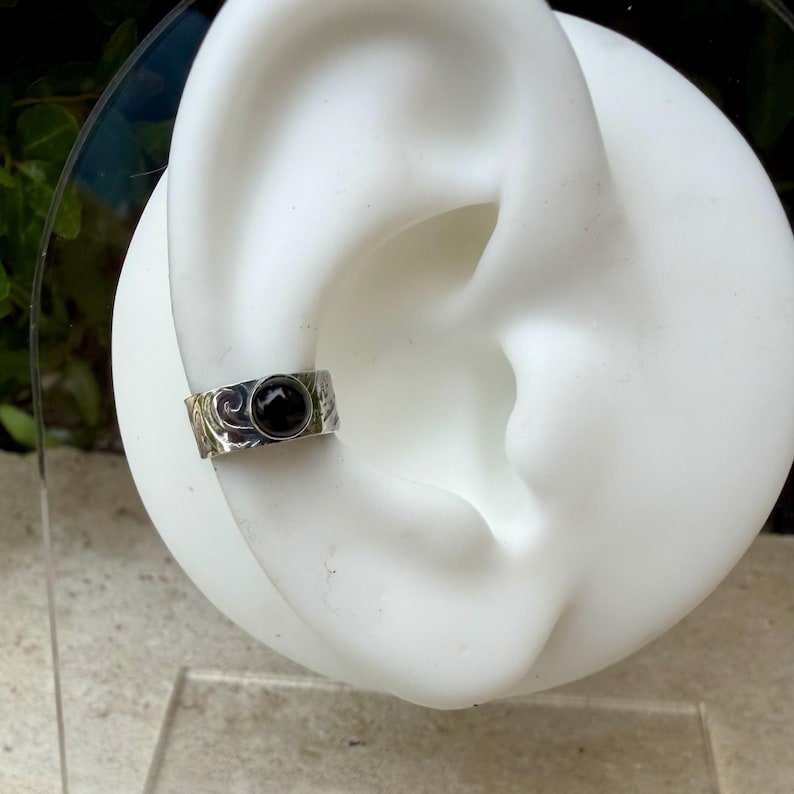 May include: A silver ear cuff with a black stone set in the center. The cuff has a textured surface with a swirling design. The jewelry is displayed on a white ear model.