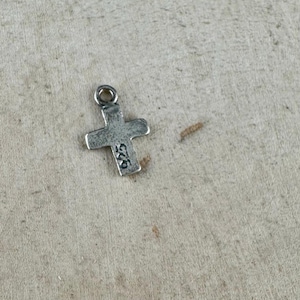 May include: A small, silver-colored cross pendant with a loop at the top for a chain. The cross has a slightly textured surface and the number "925" is stamped on one arm. The pendant is likely made of sterling silver.