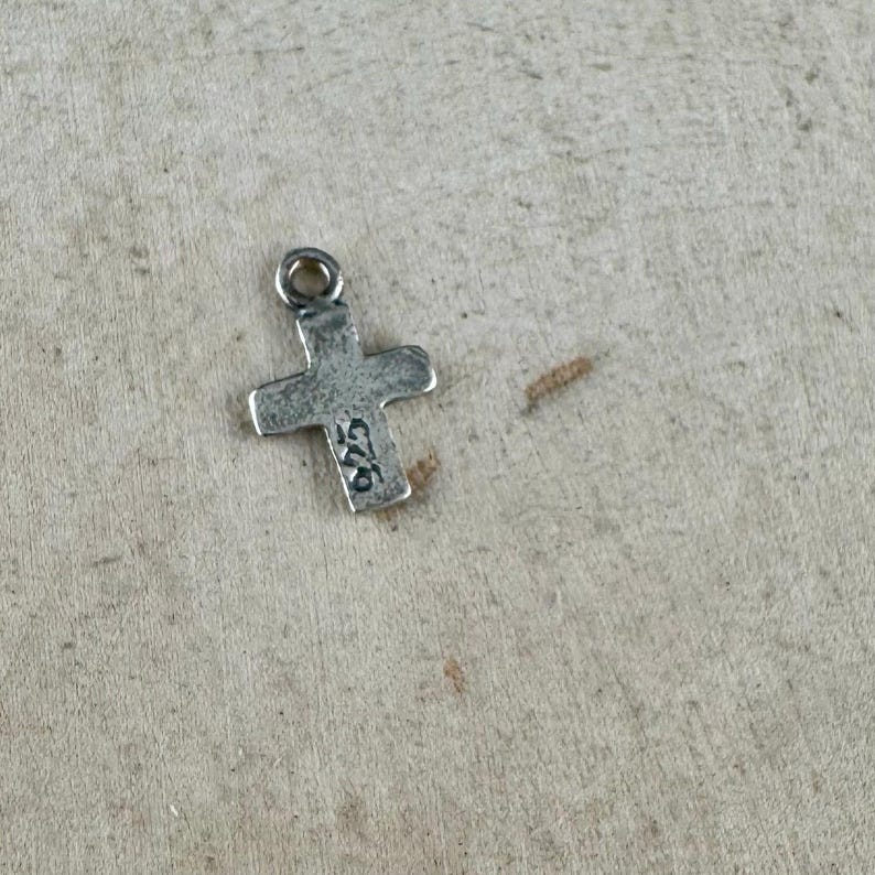 May include: A small, silver-colored cross pendant with a loop at the top for a chain. The cross has a slightly textured surface and the number "925" is stamped on one arm. The pendant is likely made of sterling silver.