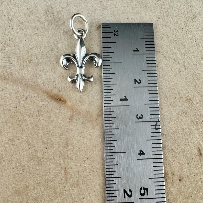 May include: A silver-colored fleur-de-lis charm with a small loop at the top. The charm is approximately 1 inch in height. A metal ruler is positioned next to the charm for scale, showing inch measurements.