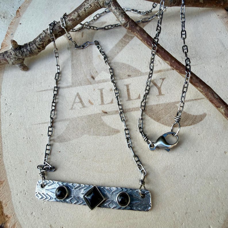 Pu&ograve; includere: Una collana color argento con un ciondolo a barra rettangolare in metallo caratterizzato da un motivo geometrico testurizzato. La barra &egrave; incastonata con due pietre nere rotonde e una pietra nera centrale a forma di diamante.