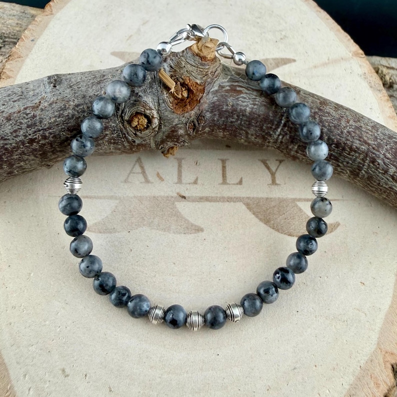 May include: A beaded bracelet featuring round, dark gray labradorite stones interspersed with small, ribbed silver-toned metal beads. The bracelet is secured with a silver-toned lobster claw clasp and a circular jump ring, creating a circular loop.