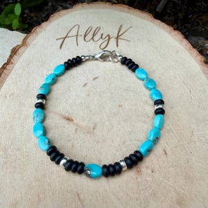May include: Turquoise and black beaded bracelet with silver accents. The bracelet features oval turquoise stones interspersed with black beads and silver spacers. The clasp is silver. The bracelet is displayed on a wooden surface, with the name "Allyk" written on the wood.