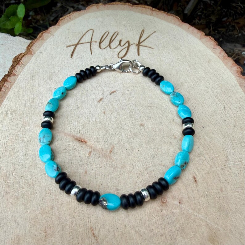 May include: Turquoise and black beaded bracelet with silver accents. The bracelet features oval turquoise stones interspersed with black beads and silver spacers. The clasp is silver. The bracelet is displayed on a wooden surface, with the name "Allyk" written on the wood.