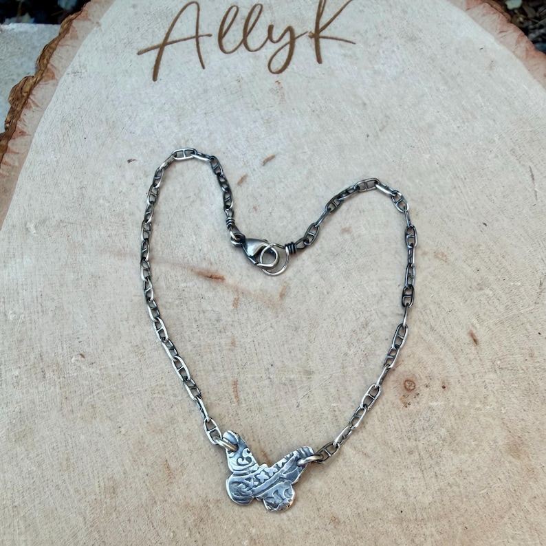 Op de afbeelding: Een zilverkleurige ketting met een vlinderhanger. De ketting heeft een schakelontwerp met een karabijnsluiting. De vlinderhanger heeft ingewikkelde details. Het woord "Allyk" staat in cursief bovenaan.