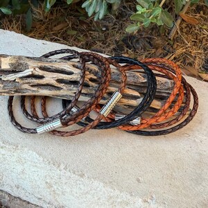 May include: A collection of braided leather bracelets in various shades of brown and black. Each bracelet features a cylindrical silver-toned metal magnetic clasp. The bracelets are arranged in a layered stack, showcasing the braided texture and the metallic finish of the closures.