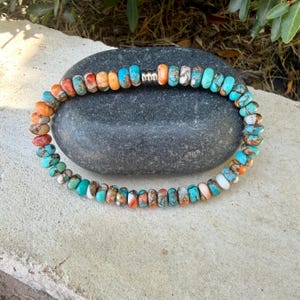 May include: A circular beaded bracelet featuring small, round, multi-colored stones in shades of turquoise, orange, white, and brown. The beads are arranged in a continuous loop, accented by a single, small, metallic silver-toned cylindrical bead.