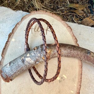 Puede incluir: Una pulsera de cord&oacute;n de cuero trenzado marr&oacute;n con un cierre met&aacute;lico cil&iacute;ndrico. La pulsera est&aacute; formada por una sola hebra de cuero tejido que se enrolla para crear una apariencia de doble capa, asegurada por una pieza de conexi&oacute;n met&aacute;lica oscura.