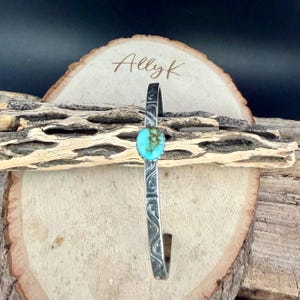 Op de afbeelding: Een zilverkleurige manchetarmband met een turquoise steen in het midden. De armband heeft een ge&euml;tst ontwerp langs de band. De armband wordt gepresenteerd op een houten oppervlak. De naam "Allyk" staat op een houten plaatje.