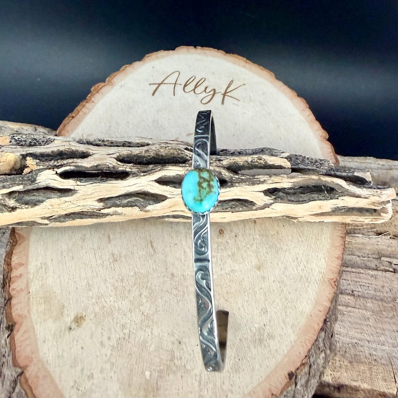 Op de afbeelding: Een zilverkleurige manchetarmband met een turquoise steen in het midden. De armband heeft een ge&euml;tst ontwerp langs de band. De armband wordt gepresenteerd op een houten oppervlak. De naam "Allyk" staat op een houten plaatje.