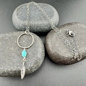 May include: A silver-toned necklace with a turquoise stone and feather pendant. The necklace features a circular design with a twisted rope texture, a small turquoise stone, and a detailed feather charm. The chain is delicate and rests on a dark gray stone.