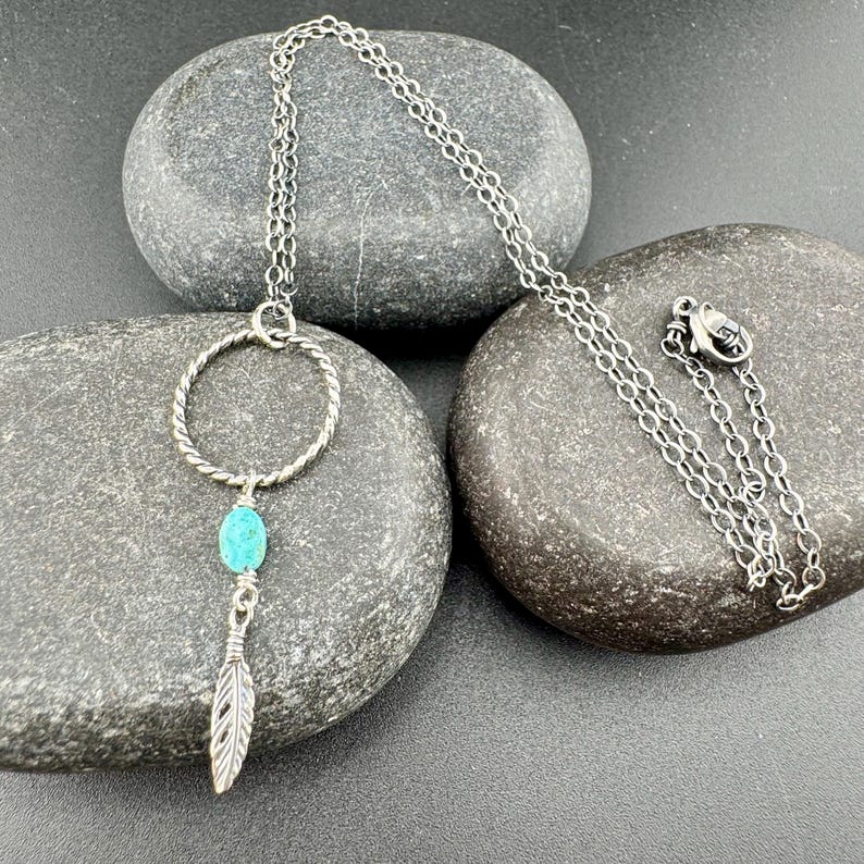 May include: A silver-toned necklace with a turquoise stone and feather pendant. The necklace features a circular design with a twisted rope texture, a small turquoise stone, and a detailed feather charm. The chain is delicate and rests on a dark gray stone.