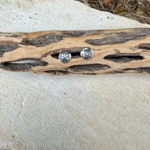 May include: A pair of circular silver stud earrings, each featuring a polished metallic surface with a recessed cross design centered on the face. The earrings are displayed side by side, showcasing the reflective finish and the detailed cross engraving.
