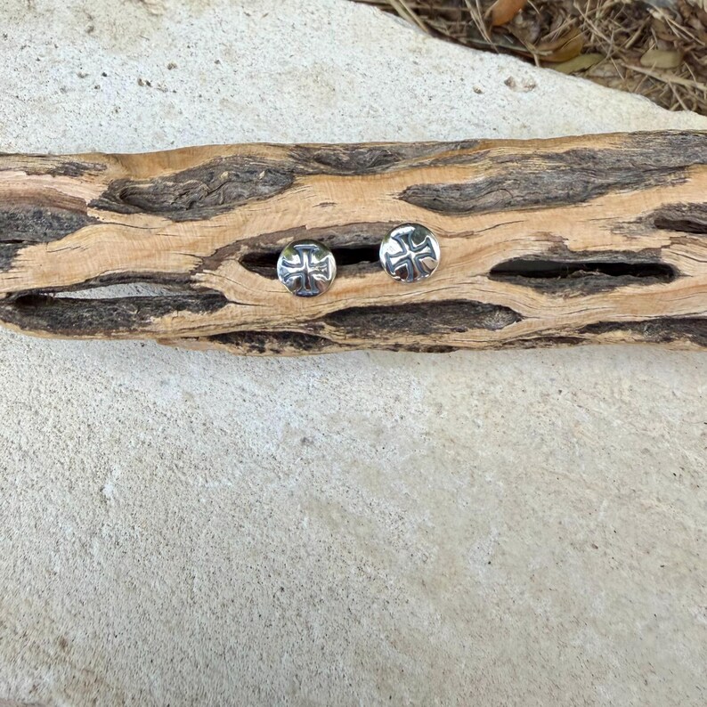May include: A pair of circular silver stud earrings, each featuring a polished metallic surface with a recessed cross design centered on the face. The earrings are displayed side by side, showcasing the reflective finish and the detailed cross engraving.
