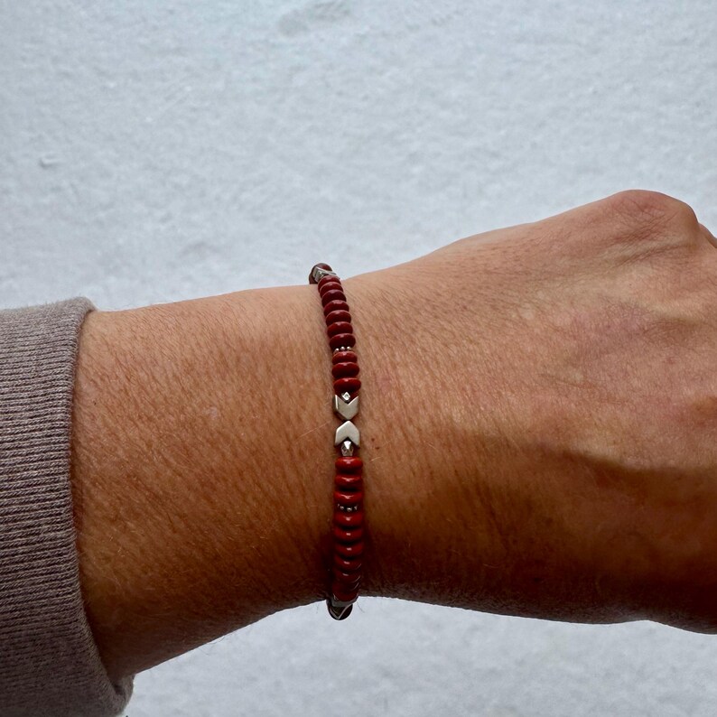 May include: Red beaded bracelet with silver-colored arrow-shaped accents. The bracelet is made of small, round, red beads strung together with silver-colored metal. The bracelet is worn on a wrist.
