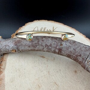 May include: A gold-colored cuff bracelet with two turquoise-colored stones. The bracelet is resting on a piece of wood. The word "Allink" is written on the bracelet.