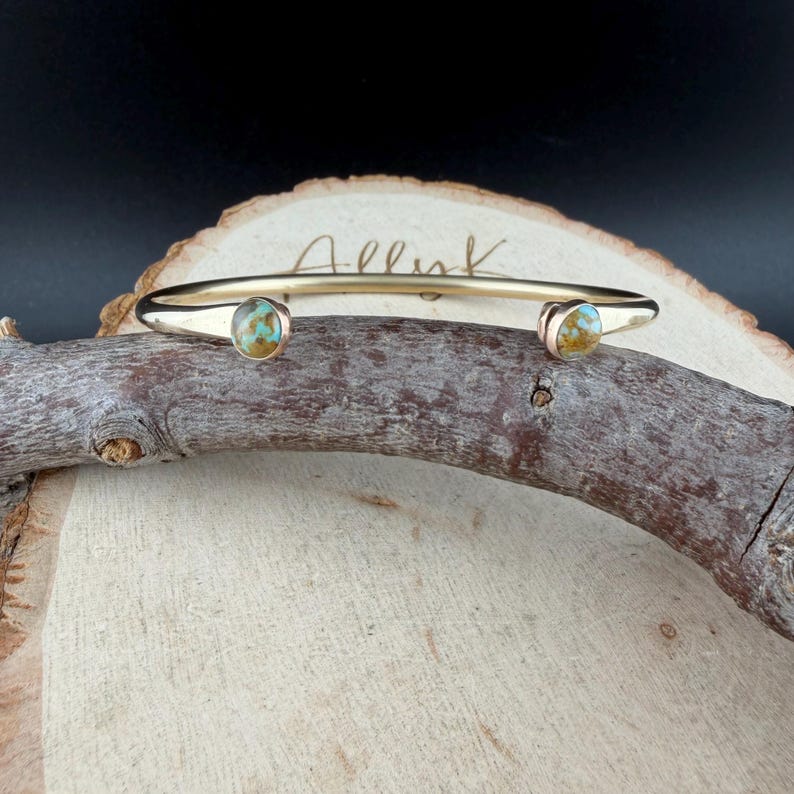 May include: A gold-colored cuff bracelet with two turquoise-colored stones. The bracelet is resting on a piece of wood. The word "Allink" is written on the bracelet.