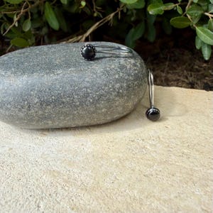 May include: A pair of silver-toned earrings featuring a single round black stone set in a polished metal frame. One earring rests horizontally on a smooth gray stone, while the other hangs vertically from the side of the stone surface.