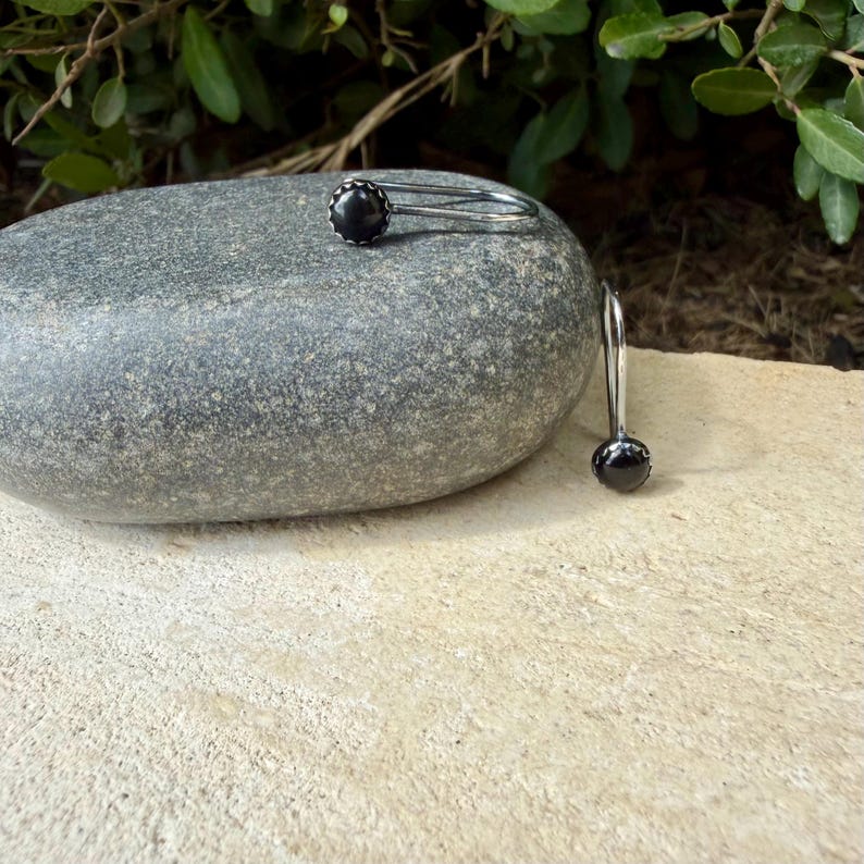 May include: A pair of silver-toned earrings featuring a single round black stone set in a polished metal frame. One earring rests horizontally on a smooth gray stone, while the other hangs vertically from the side of the stone surface.