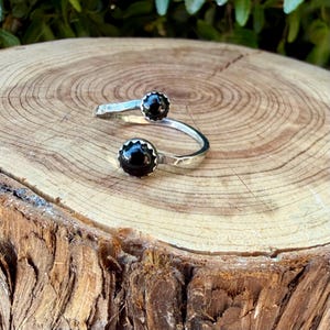 May include: A silver bypass ring featuring two round, polished black stones set in scalloped metal bezels. The band has a textured, hammered finish and an open design, allowing the two ends to overlap slightly while showcasing the dark stones.