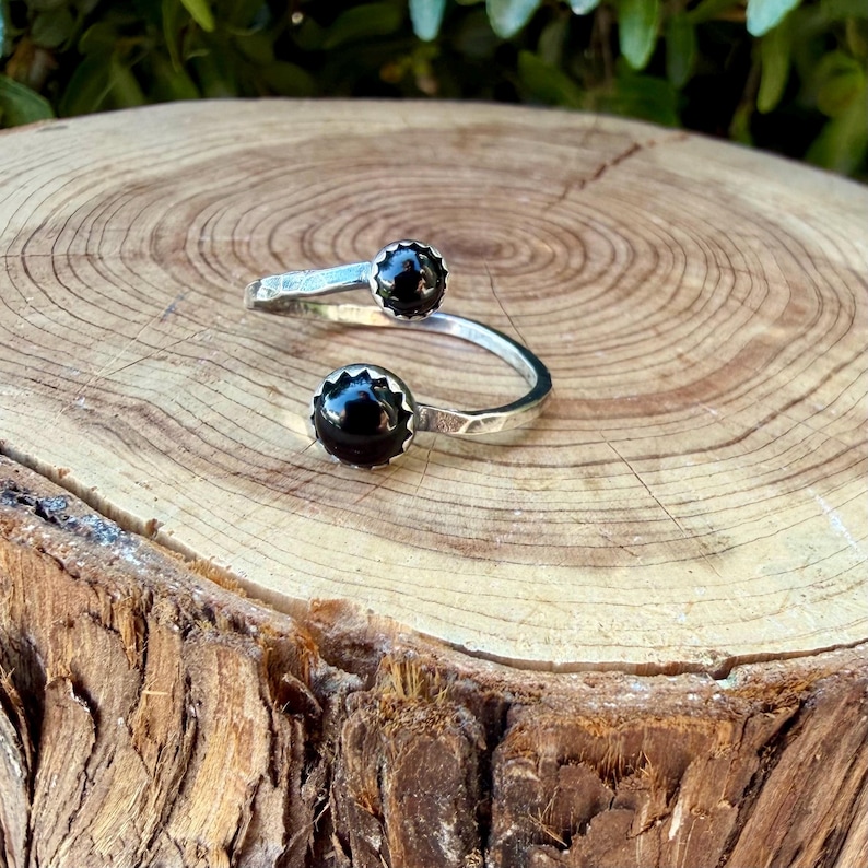May include: A silver bypass ring featuring two round, polished black stones set in scalloped metal bezels. The band has a textured, hammered finish and an open design, allowing the two ends to overlap slightly while showcasing the dark stones.