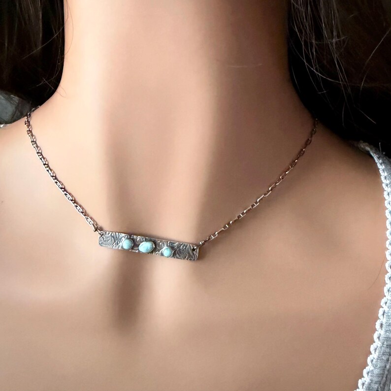 May include: A silver-toned necklace featuring a rectangular bar pendant with an embossed floral pattern and three small, round, light blue stones set horizontally across the center, suspended from a delicate silver-toned chain.
