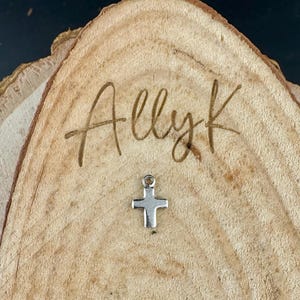 May include: A small, silver-colored cross charm with a loop at the top. The charm is set on a wooden surface with the engraved text "Allyk". The wood has a natural, light brown color and visible grain.